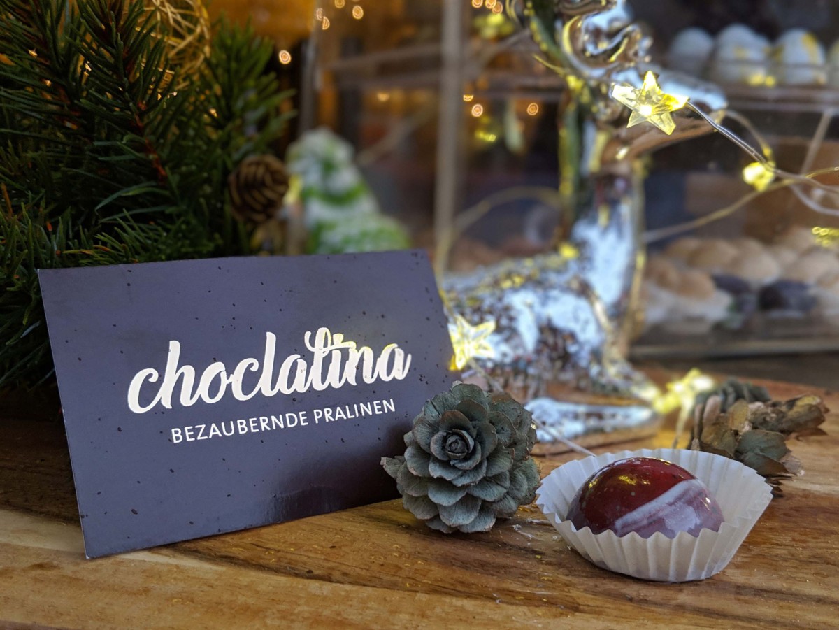 Eine schwarze Karte auf der "choclatina" in Goldschrift steht, lehnt an einen Tannenzweig auf einem Holztisch. Vor der Karte liegen weihnachtliche Dekorationen und eine rote Praline.
