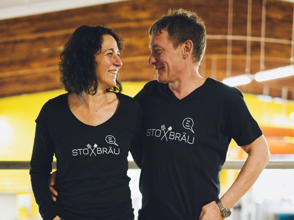 Ein Mann und eine Frau tragen ein Shirt mit der Aufschrift Stoxbräu und lachen sich an.