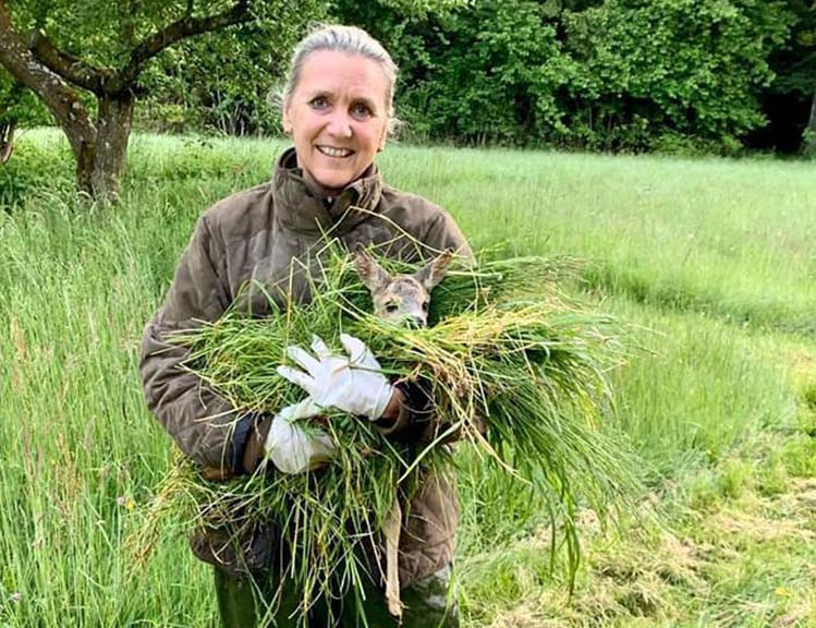 Frau im Jägeroutfit hält ein gerettetes, in Gras gebettetes Kitz in den Armen