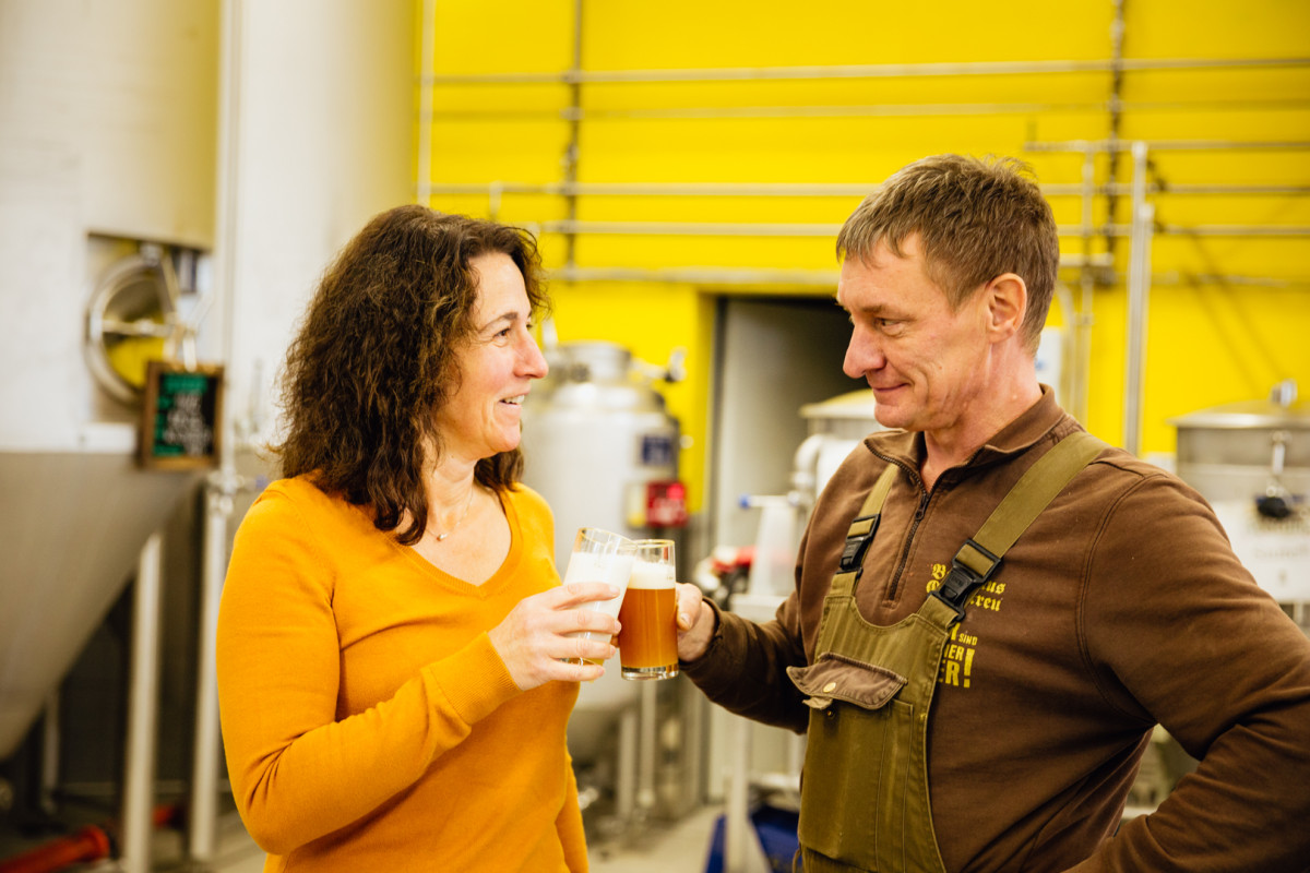 Eine Frau und ein Mann stehet vor einer gelben Wand und stoßen mit einem Glas Bier an. Sie sehen sich an und lächeln.